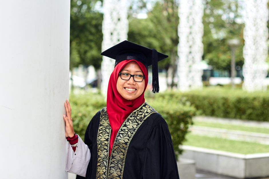 A smiling Southeast Asian woman in cap and gown celebrating graduation outdoors in Putrajaya.